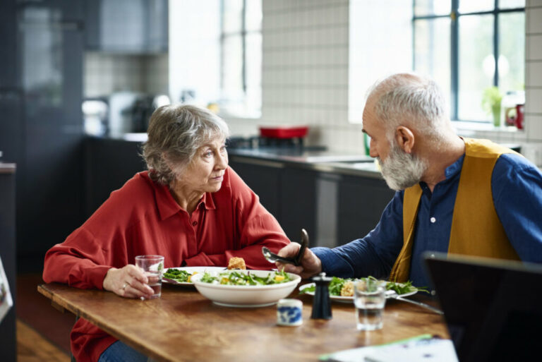 Retired man and woman sitting at dining table in modern kitchen, eating salad and talking, woman looking at man and listening with surprised expression